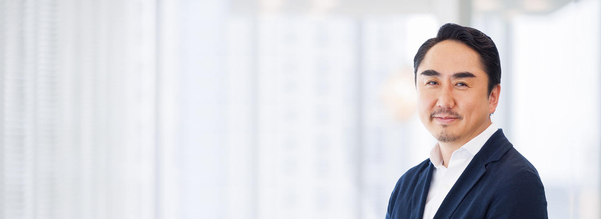 This is a photo of CEO Takeshi Idezawa. A man in a suit is smiling in a bright and modern office. There is a large window in the background.