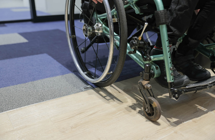 A person in a wheelchair attempting to cross the boundary between carpet and wooden flooring with the front wheels. The foot and wheelchair frame are visible.