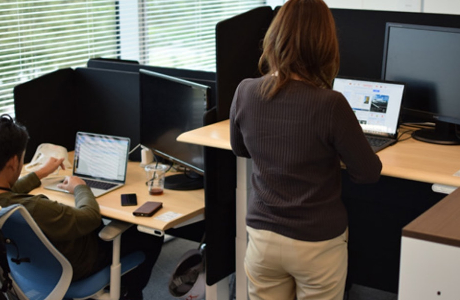A woman in the foreground operating a laptop on an electric adjustable desk, while a man in the background sits and operates a laptop.