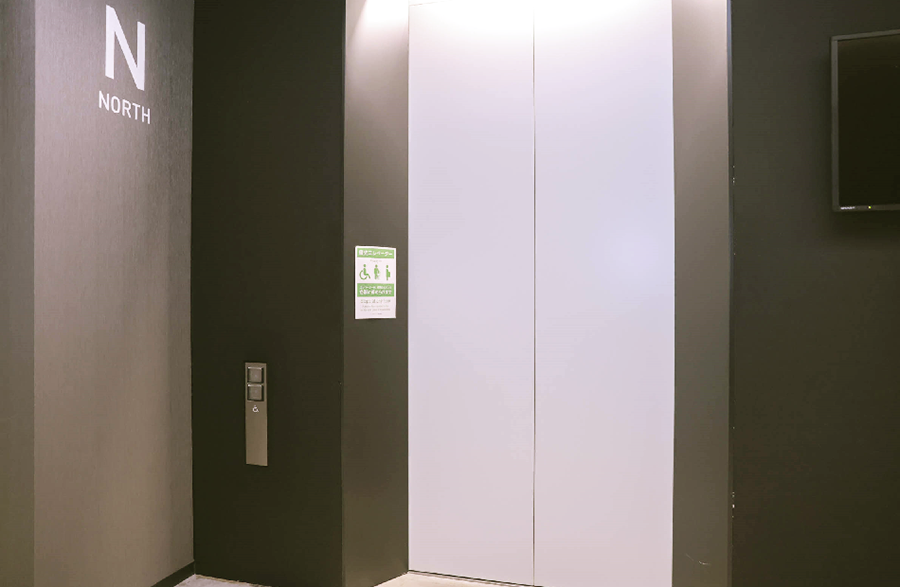 An elevator with a white door next to a black wall labeled "N NORTH," with a wheelchair button and green explanatory signage attached.