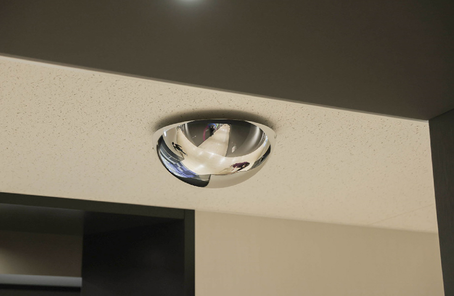 A hemispherical convex mirror mounted on the ceiling, reflecting surrounding corridors and parts of people.