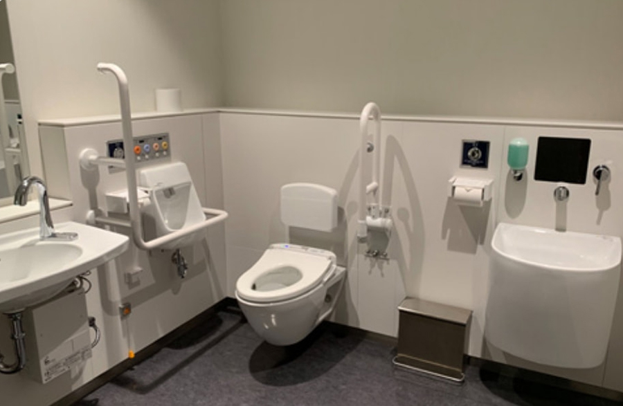 Interior of a barrier-free multipurpose restroom. Equipped with a Western-style toilet, multiple handrails, a sink, a soap dispenser, an emergency button, and a diaper changing table.