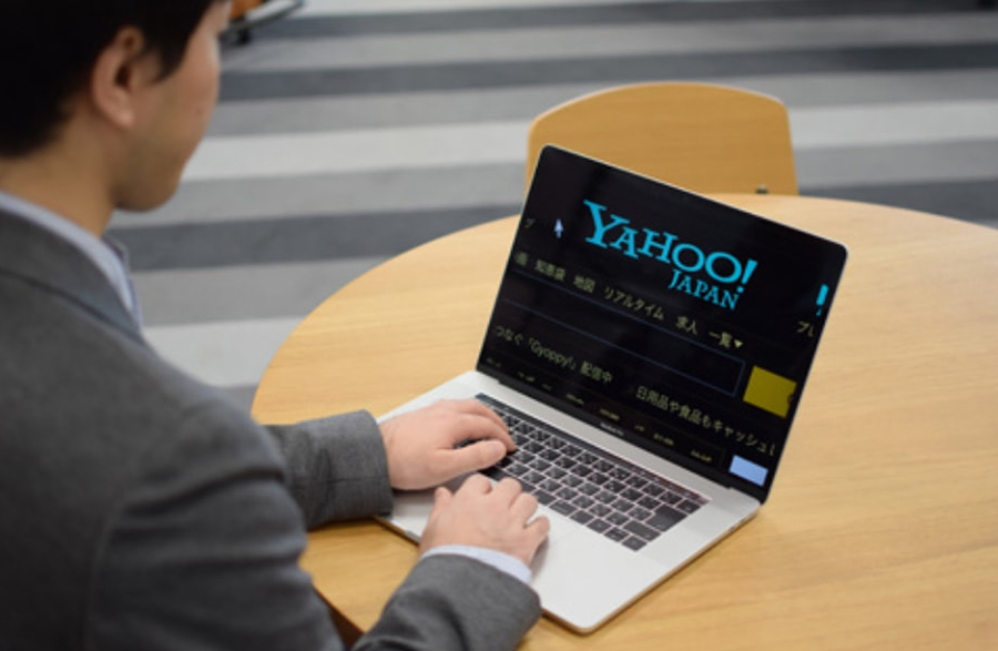 A man sitting at a round wooden table, operating a laptop. The screen prominently displays Yahoo! JAPAN's top page with a black background, reflecting a color setting change.
