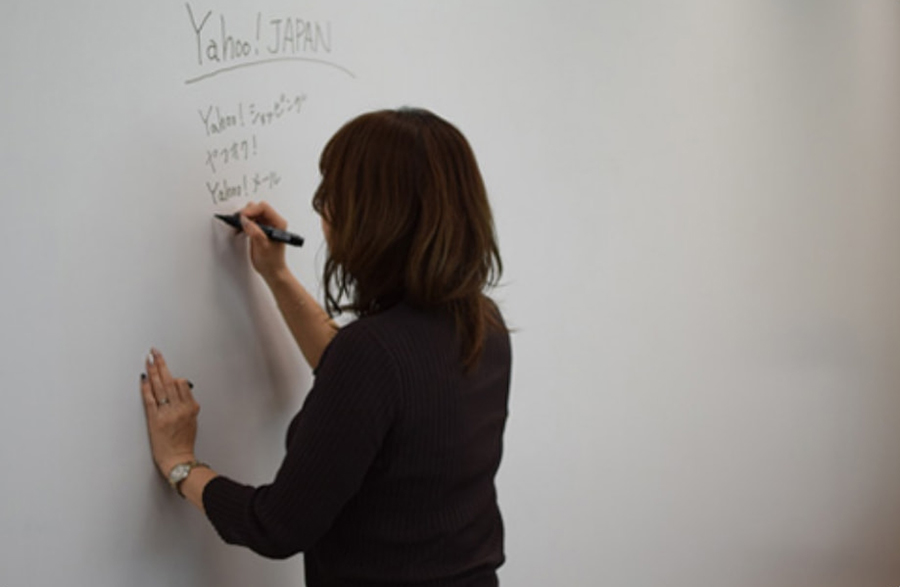 A woman standing in front of a whiteboard covering an entire wall, writing "Yahoo! JAPAN" and "Shopping" with a black marker.